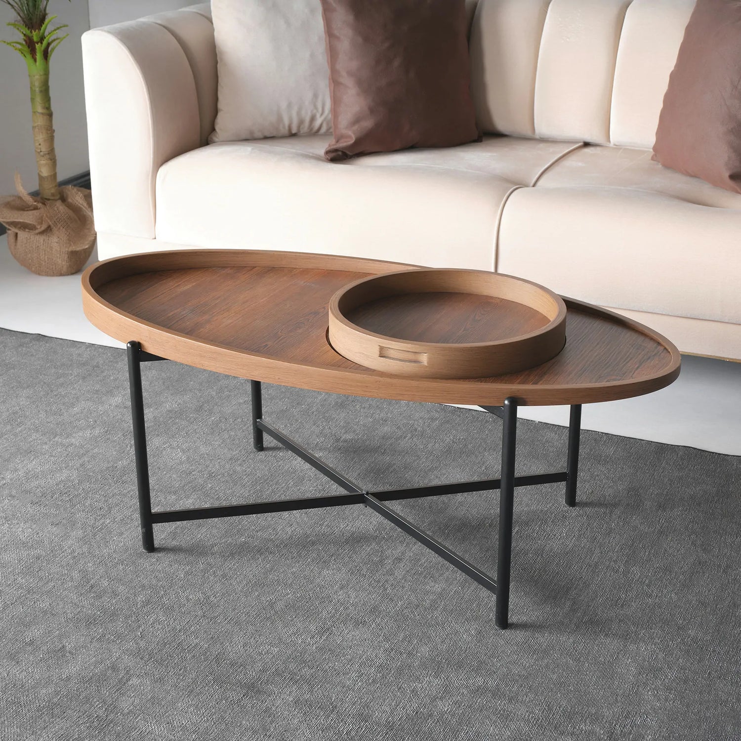 An oval-shaped coffee table with a natural wood finish and black metal legs, featuring a removable tray. The table is placed on a grey carpet, with a beige sofa in the background. By Papella furniture design