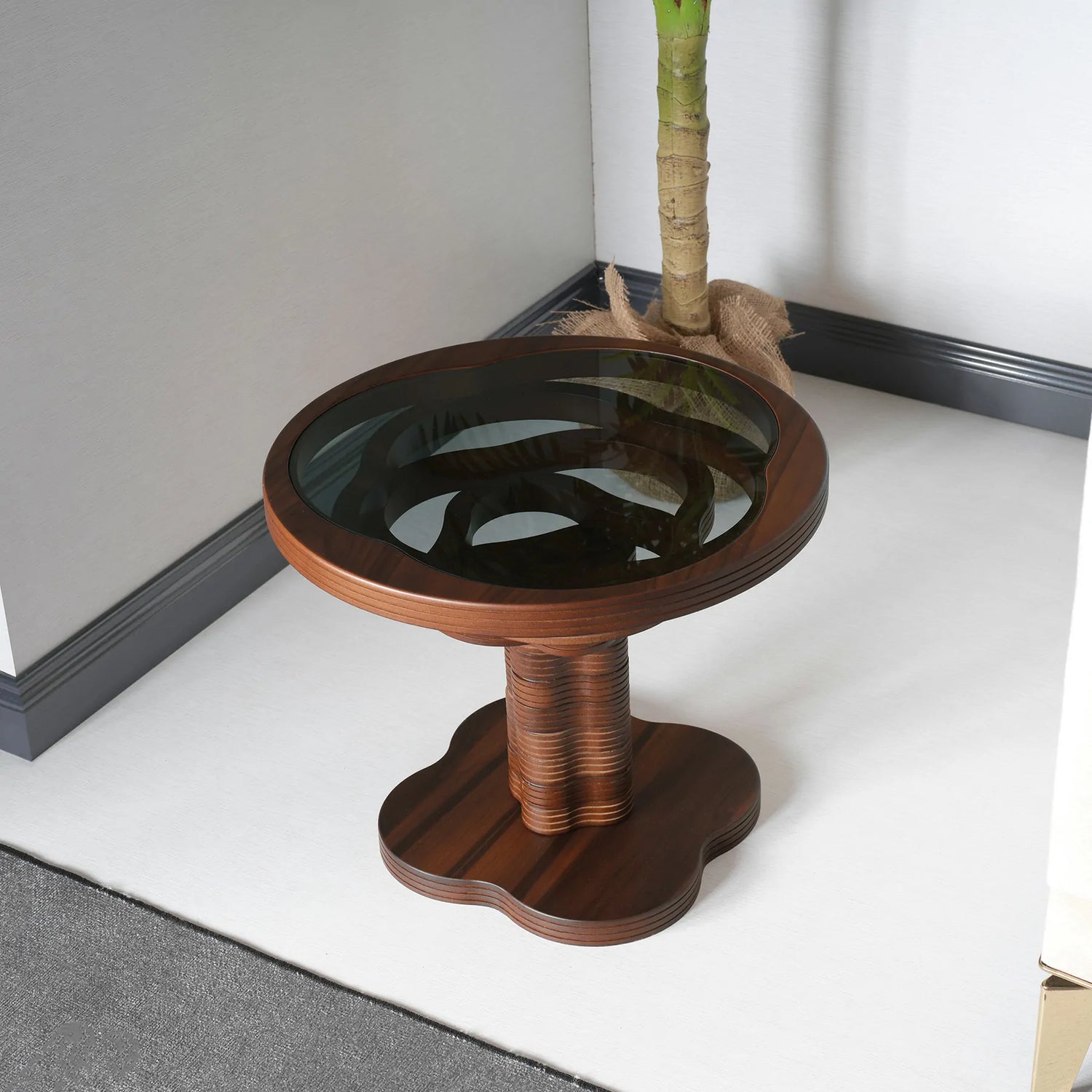 A side table with a 3D wood design base and a dark glass top, placed on a white floor against a wall.