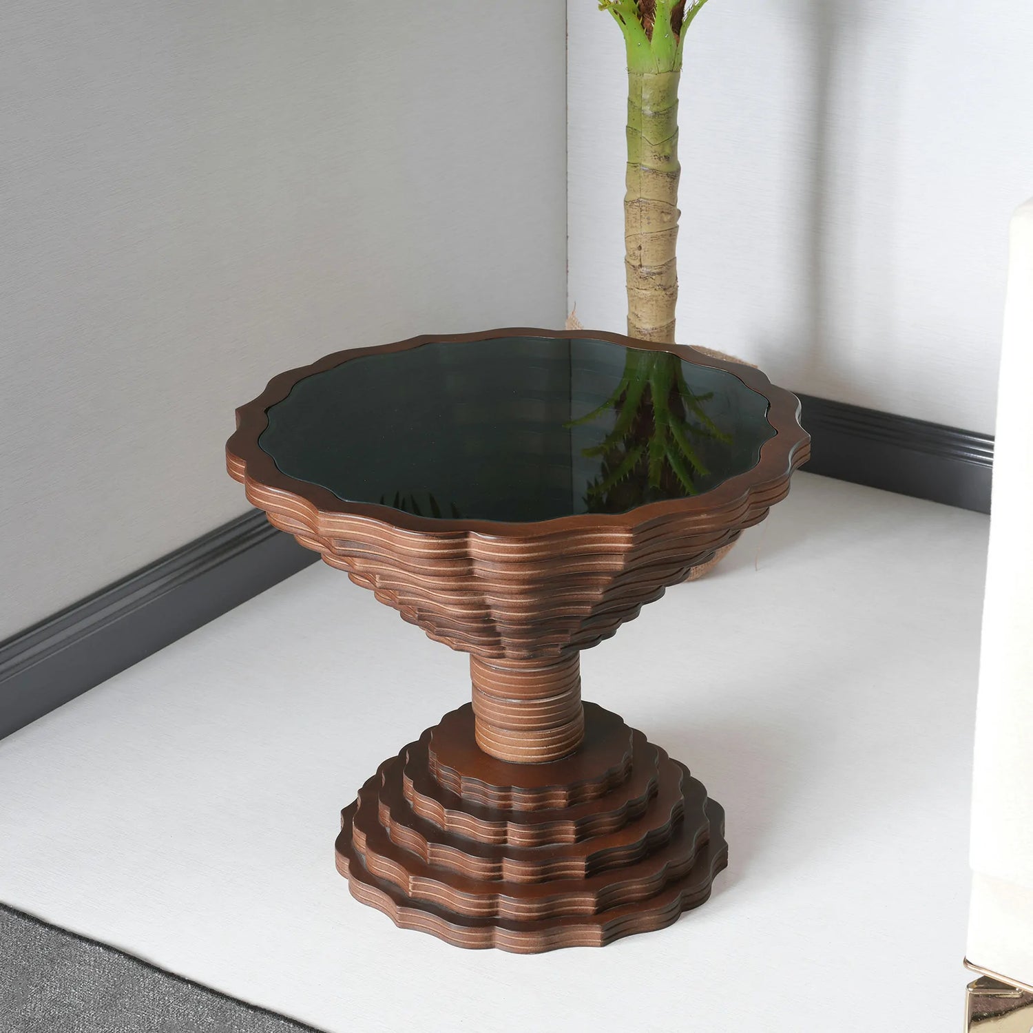 A round wooden side table with a scalloped edge design and a dark glass top, placed on a white floor.