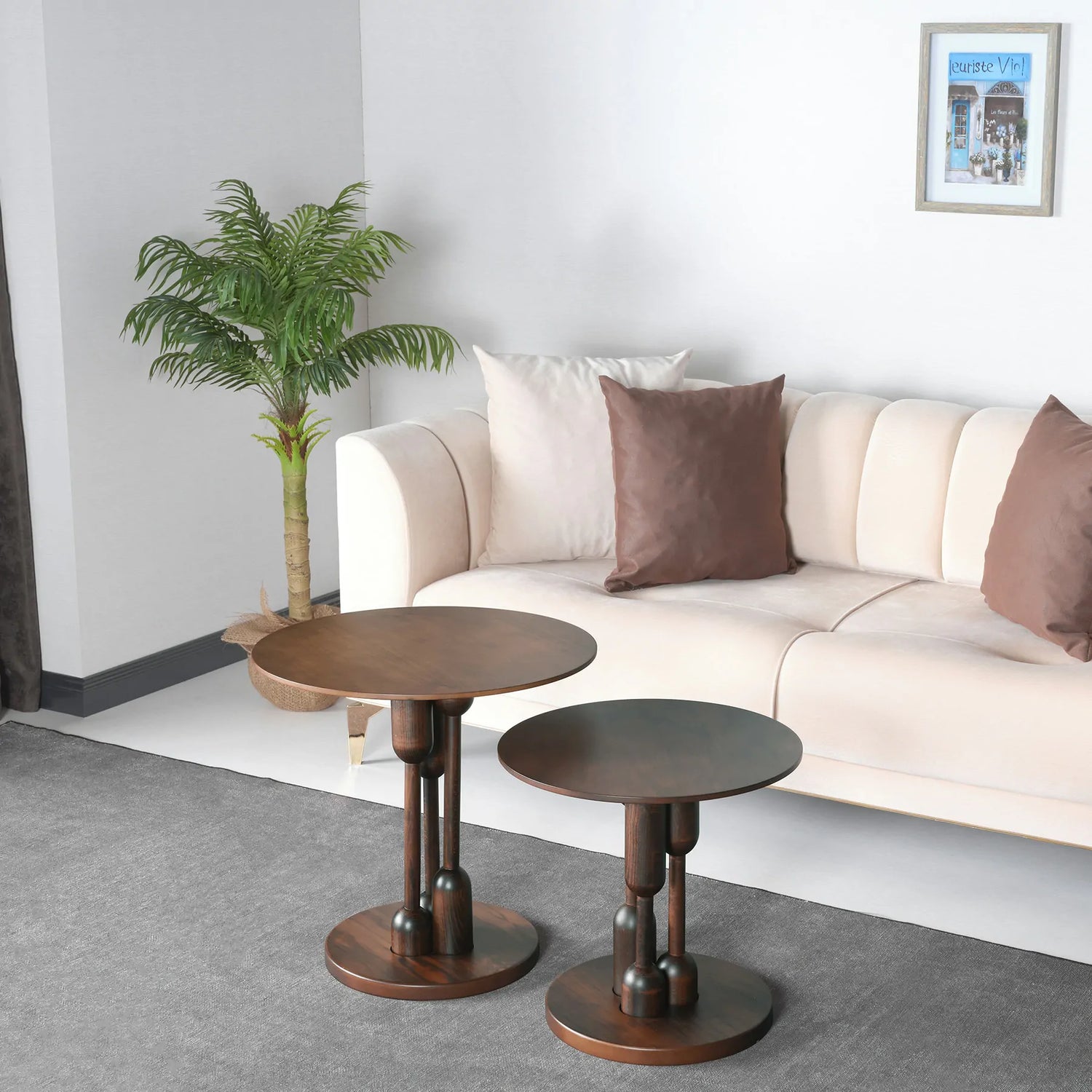 Two round wooden side tables with a twin design, one larger than the other, placed on a grey floor in front of a beige sofa with brown and white cushions. There is a green plant to the left of the larger table and a framed picture on the wall above the sofa.