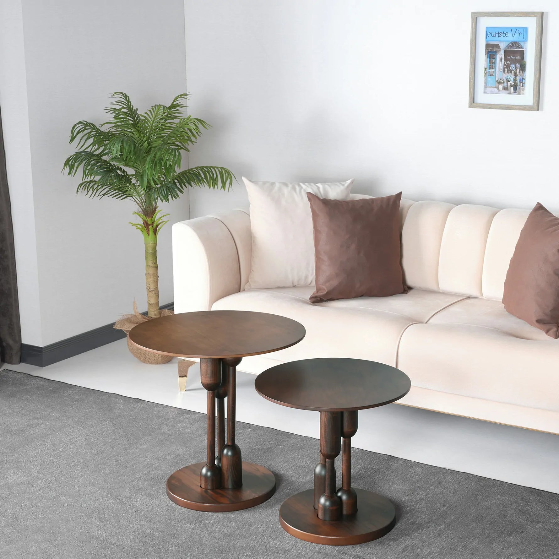 Two round wooden side tables with a twin design, one larger than the other, placed on a grey floor in front of a beige sofa with brown and white cushions. There is a green plant to the left of the larger table and a framed picture on the wall above the sofa.