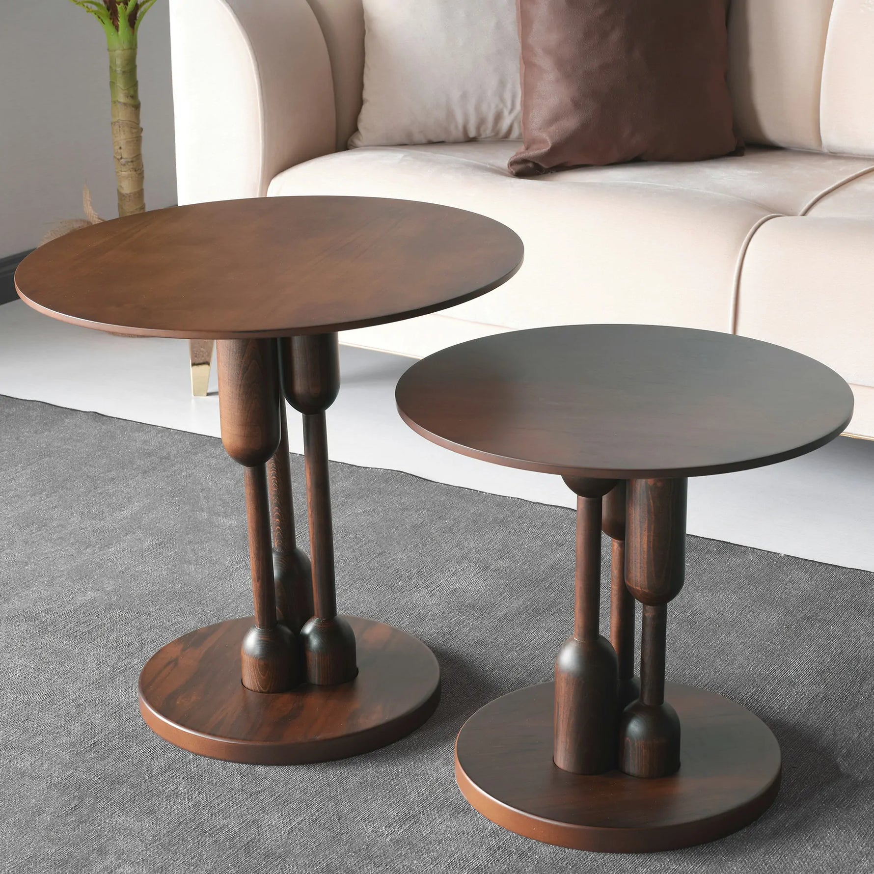 A stylish round coffee table with a dark wood finish and distinctive designed legs, placed on a grey carpet in a room with a beige sofa and a potted plant in the background.