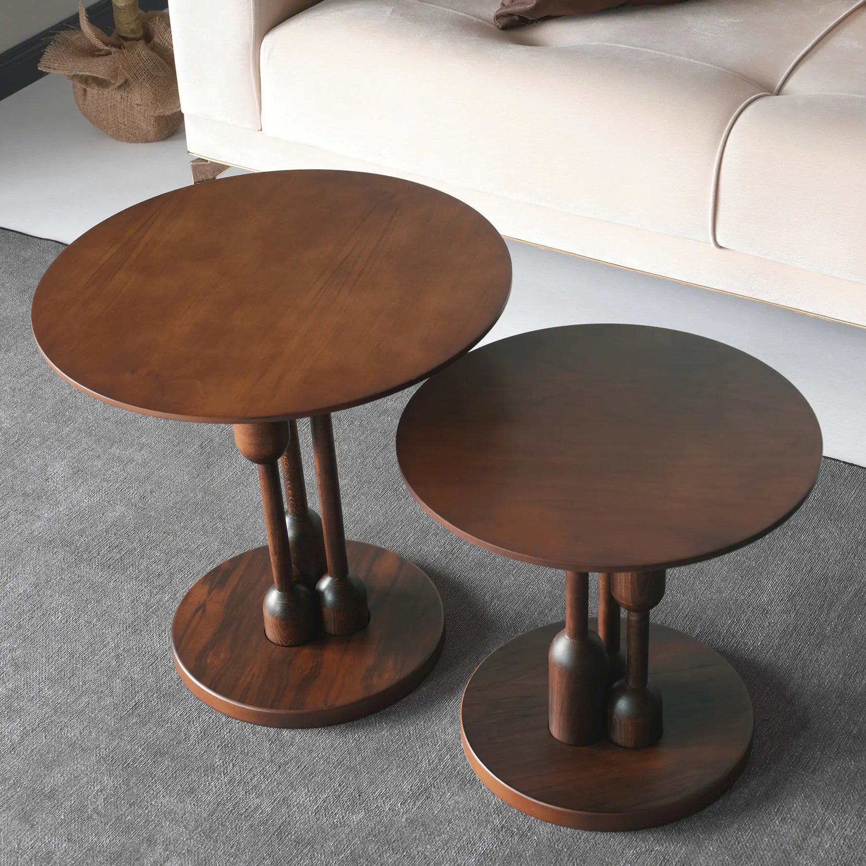 A stylish round coffee table with a dark wood finish and distinctive designed legs, placed on a grey carpet in a room with a beige sofa and a potted plant in the background.