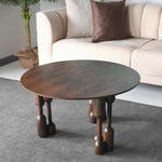 A stylish round coffee table with a dark wood finish and distinctive designed legs, placed on a grey carpet in a room with a beige sofa and a potted plant in the background.