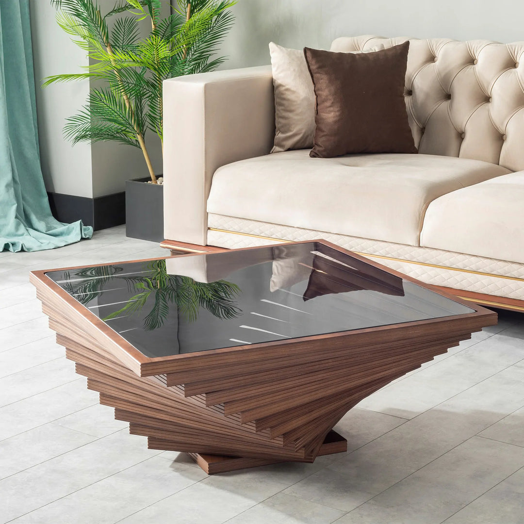 A square-shaped coffee table with a twisted design in the wood structure, featuring a dark glass top, placed on a tiled floor next to a beige sofa with a brown cushion.

