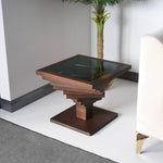 A square-shaped coffee table with a twisted design in the wood structure, featuring a dark glass top, placed on a tiled floor next to a beige sofa with a brown cushion.

