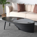 An oval-shaped coffee table with a layered walnut base and a tempered glass top, placed in a room with a sofa and a plant in the background.